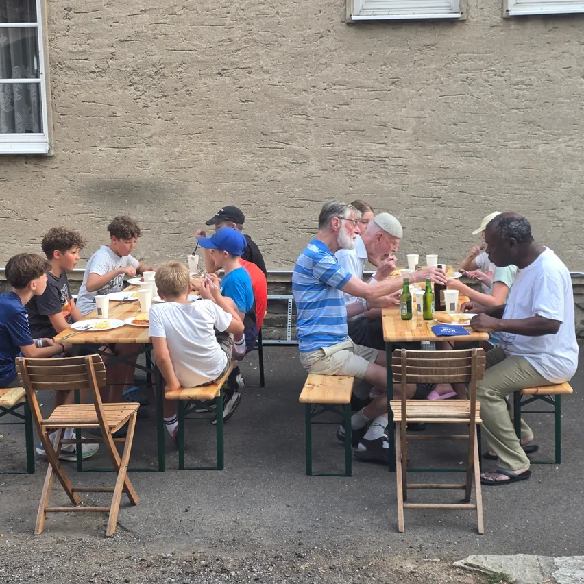 Eine Gruppe von Menschen, einschließlich Kinder und Erwachsene, sitzen draußen an Tischen, wahrscheinlich in einem Park oder Garten. Sie essen und trinken aus Tassen und Flaschen.