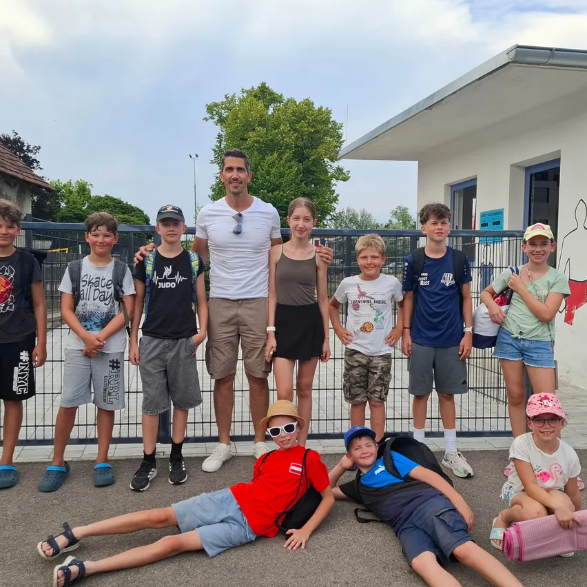 Eine Gruppe von Kindern und ein erwachsener Mann posieren für ein Foto vor einem Gebäude. Die Kinder tragen Freizeitkleidung und einige tragen Hüte. Der erwachsene Mann trägt ein weißes Hemd und eine Sonnenbrille.