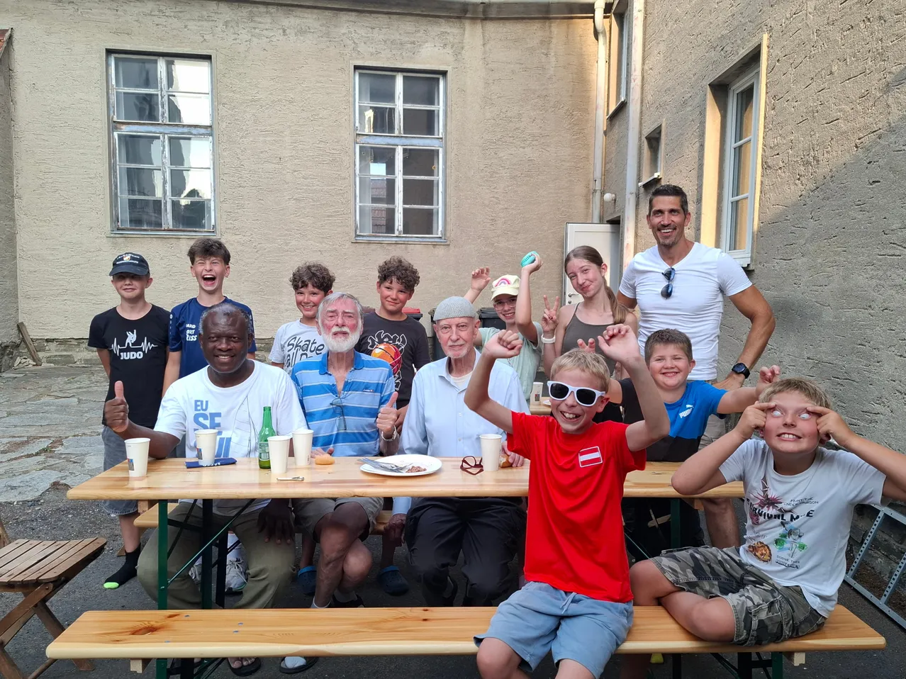 Eine Gruppe von Menschen, einschließlich Erwachsener und Kindern, versammelt sich um einen Tisch im Freien. Sie lächeln und posieren für ein Foto. Der Tisch hat Tassen, Teller und Flaschen. Hinter ihnen steht ein Gebäude mit Glasfenstern.