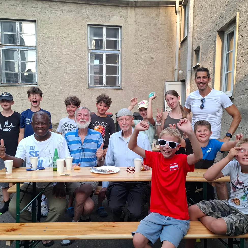 Eine Gruppe von Menschen, einschließlich Erwachsener und Kindern, versammelt sich um einen Tisch im Freien. Sie lächeln und posieren für ein Foto. Der Tisch hat Tassen, Teller und Flaschen. Hinter ihnen steht ein Gebäude mit Glasfenstern.