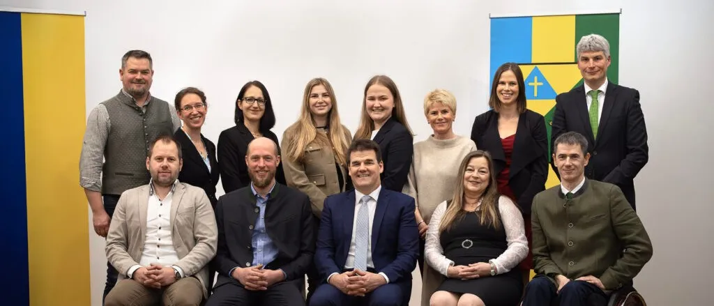 Eine Gruppe von Menschen posiert für ein Foto. Sie sitzen in einer Reihe auf Stühlen. Sie alle lächeln und scheinen für ein Foto zu posieren. Hinter ihnen ist eine weiße Wand mit einem farbenfrohen Design.