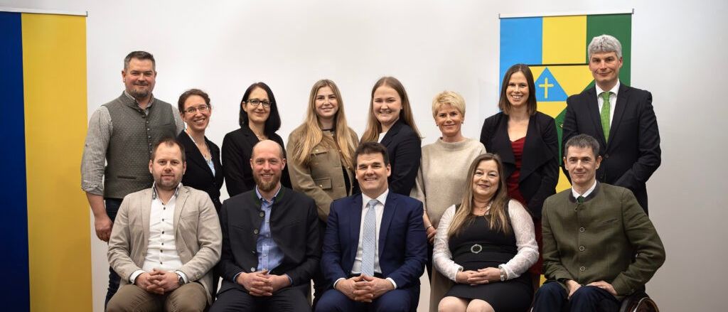 Eine Gruppe von Menschen posiert für ein Foto. Sie sitzen in einer Reihe auf Stühlen. Sie alle lächeln und scheinen für ein Foto zu posieren. Hinter ihnen ist eine weiße Wand mit einem farbenfrohen Design.