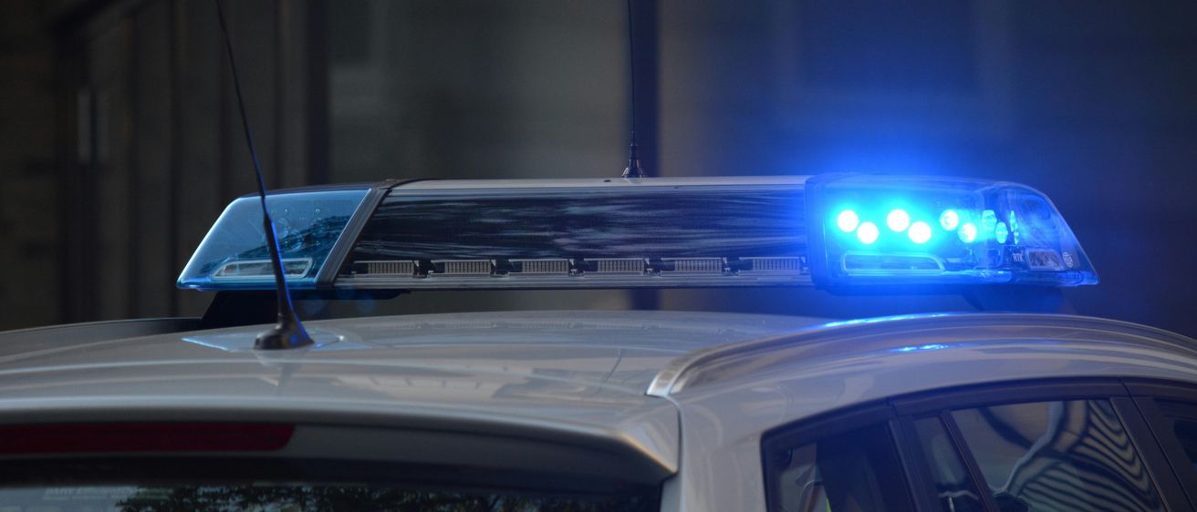 Ein weißer Wagen mit blauen Notfallleuchten auf dem Dach ist in einem dunklen Bereich geparkt, wahrscheinlich für einen Polizeieinsatz.