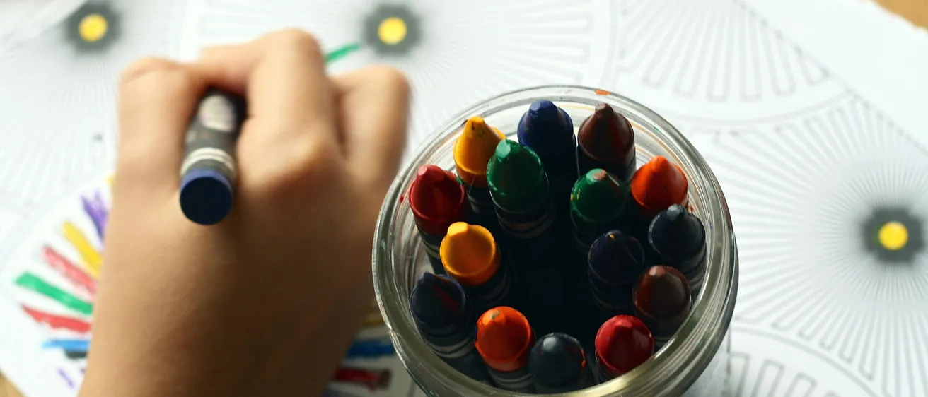 Eine Kinderhand hält einen Buntstift und färbt eine Blume auf einer Ausmalseite ein. Ein Glas mit Buntstiften ist in der Nähe, verschiedene Farben sind sichtbar.