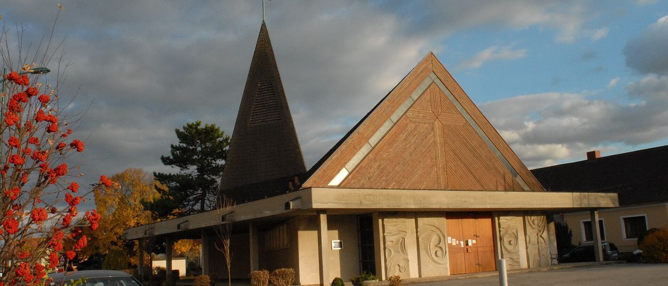 Eine moderne Kirche mit einem dreieckigen Turm und einem Kreuz darauf. Sie hat ein dreieckiges Dach und ist von einem Rasenbereich mit Pflanzen umgeben.