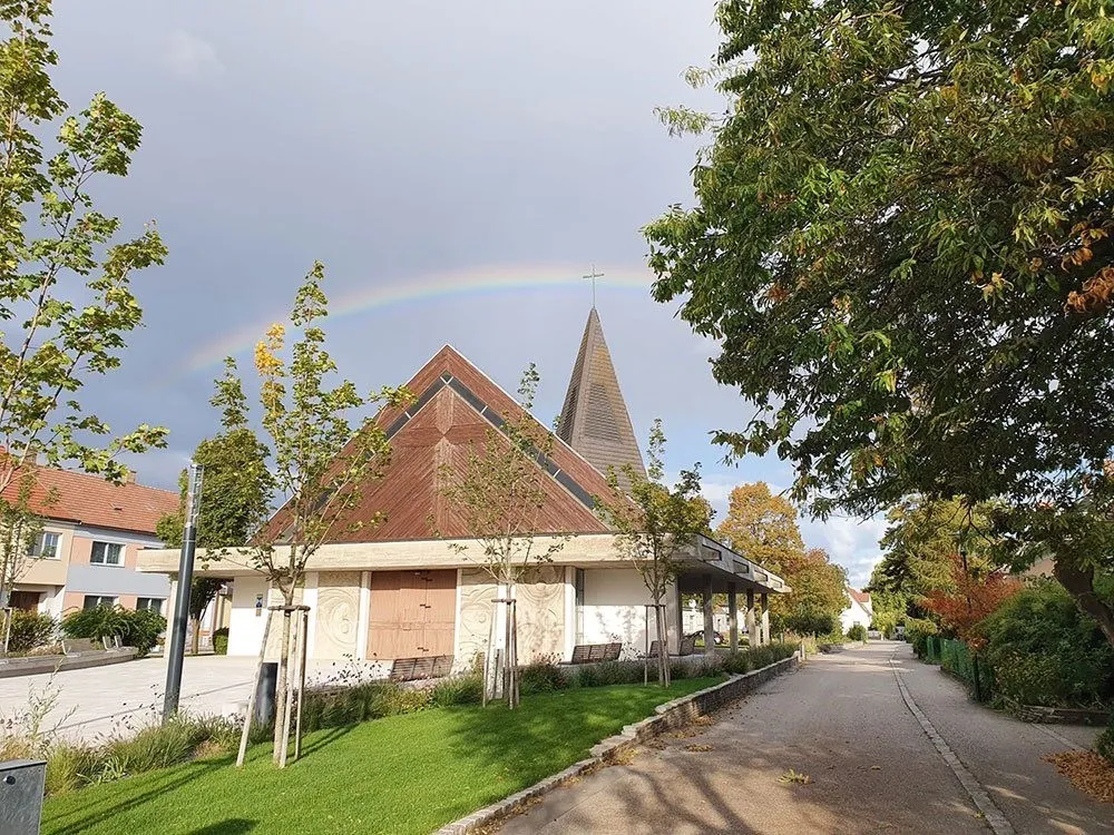 Eine Kirche mit einem Regenbogen am Himmel darüber. Sie hat ein braunes Dach und einen Turm mit einem Kreuz oben.