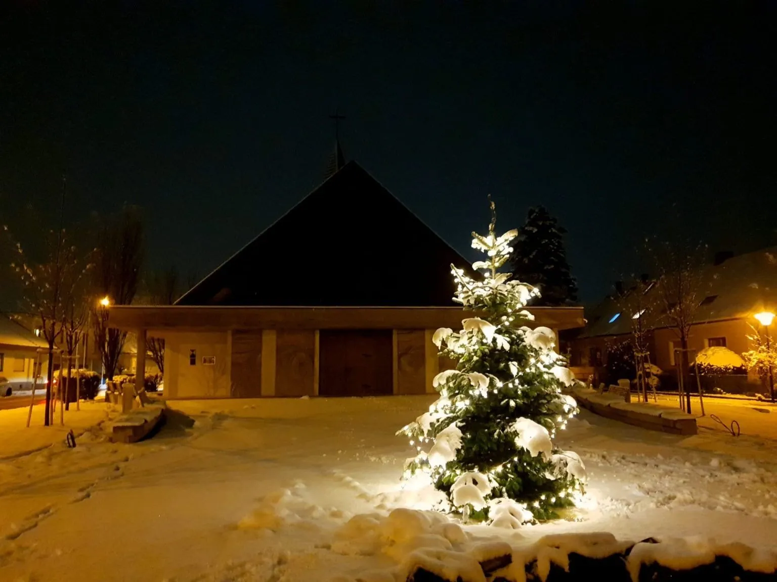 Ein beleuchteter Weihnachtsbaum steht in der Nacht im Schnee vor einer Kirche.