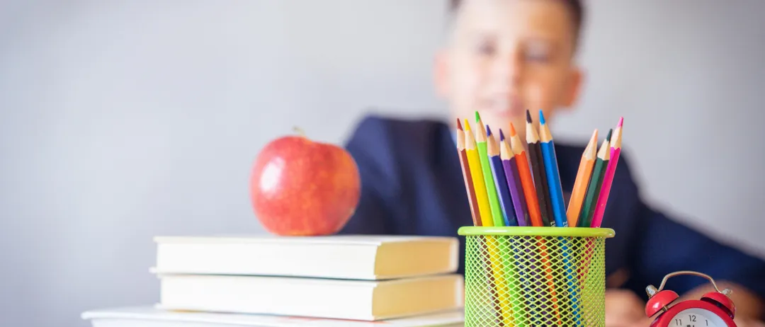 Ein Junge sitzt an einem Schreibtisch mit einem Apfel, Büchern und einem Stifthalter voller bunter Stifte. Er trägt ein blaues Hemd.