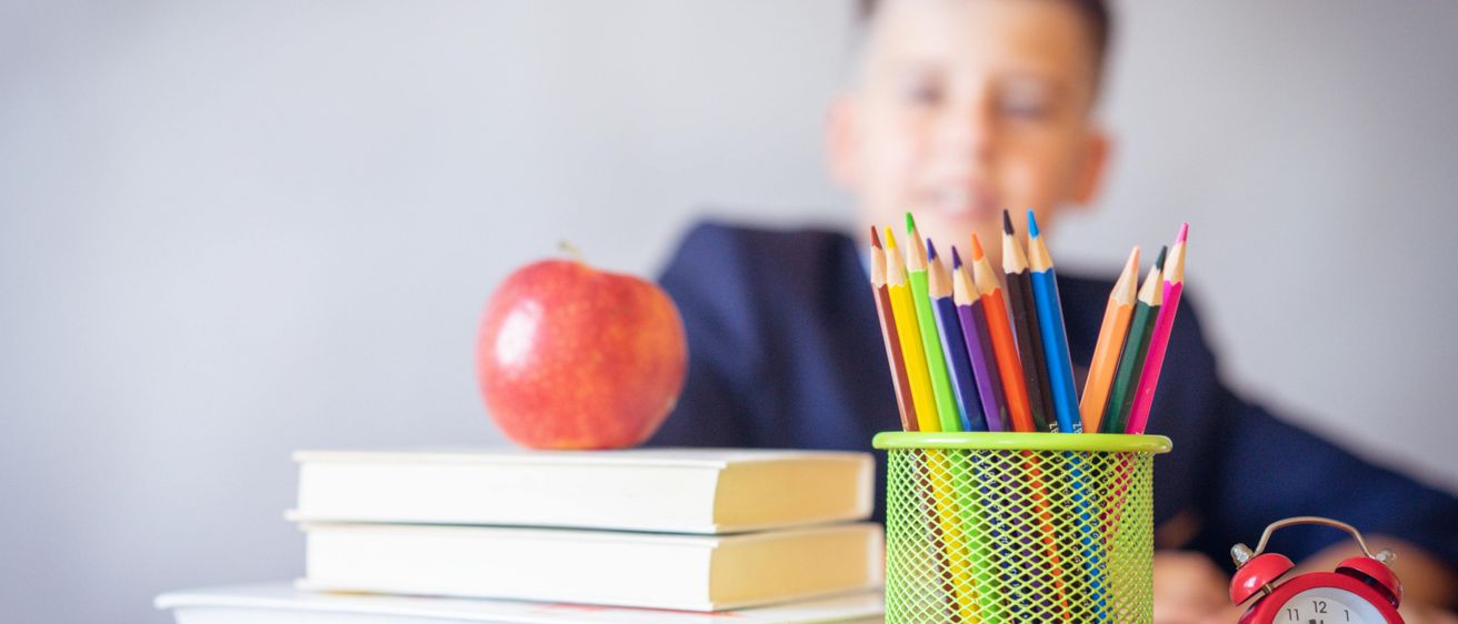 Ein Junge sitzt an einem Schreibtisch mit einem Apfel, Büchern und einem Stifthalter voller bunter Stifte. Er trägt ein blaues Hemd.