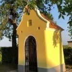 Ein kleines, gelbes Gebäude mit einem braunen Dach und einem schwarzen Tor, umgeben von einer Hecke und Bäumen, unter einem klaren blauen Himmel.