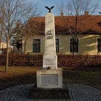 Ein Denkmal vor einem gelben Gebäude mit einem braunen Dach und Fenstern. Das Denkmal hat eine Adlerstatue auf der Spitze und Text in einer fremden Sprache.