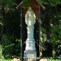 Eine Statue der Jungfrau Maria steht unter einem Baldachin in einem bewaldeten Gebiet. Die Statue steht auf einem weißen Sockel, umgeben von Metallstützen. Am Fuße befinden sich Blumen.