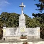Ein Marmormonument mit einem Kreuz an der Spitze, umgeben von Grün und Bäumen. Auf dem Monument ist der Name 'ROCHOLI' und darunter Text eingraviert. Das Monument steht auf einem Betonsockel und befindet sich im Freien.