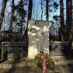 Ein Gedenkstein mit einer geätzten Figur und zwei Tafeln steht in einem Wald, mit Blumen und Grünpflanzen darum herum. Hohe Bäume umgeben das Gebiet, und der Himmel ist klar.