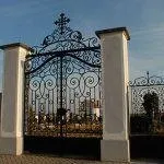 Ein schwarzes Tor aus Schmiedeeisen mit kunstvollen Verzierungen steht zwischen zwei weißen Säulen. Das Tor ist geschlossen und oben befindet sich ein Kreuz. Der Himmel ist klar mit ein paar Wolken.