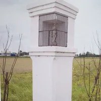 Ein weißer Pfeiler mit vergittertem Fenster steht in einem offenen Feld, umgeben von kahlen Bäumen. In der Ferne sind Gebäude und Bäume unter einem bewölkten Himmel zu sehen.