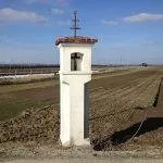 Ein weißer Glockenturm mit einem Kreuz auf der Spitze steht in der Mitte eines großen Feldes unter einem blauen Himmel mit Wolken.