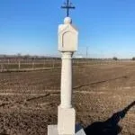 Ein weißer Grabstein mit einem Kreuz darauf steht auf einem kahlen Feld unter einem klaren Himmel. Der Grabstein ist von einem breiten, leeren Feld umgeben.