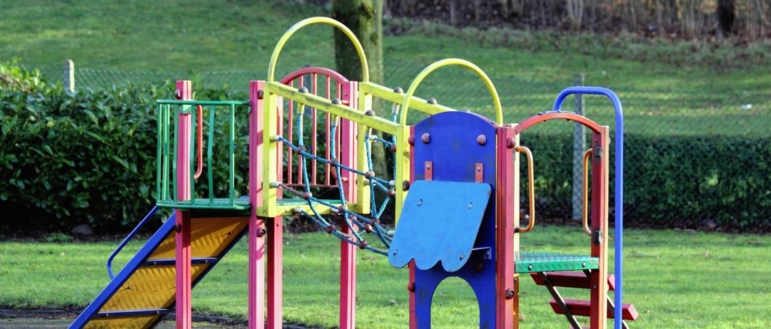 Ein farbenfroher Spielplatz mit Rutsche, Kletternetz und Treppen, auf einer Rasenfläche mit einer Hecke und Bäumen im Hintergrund.