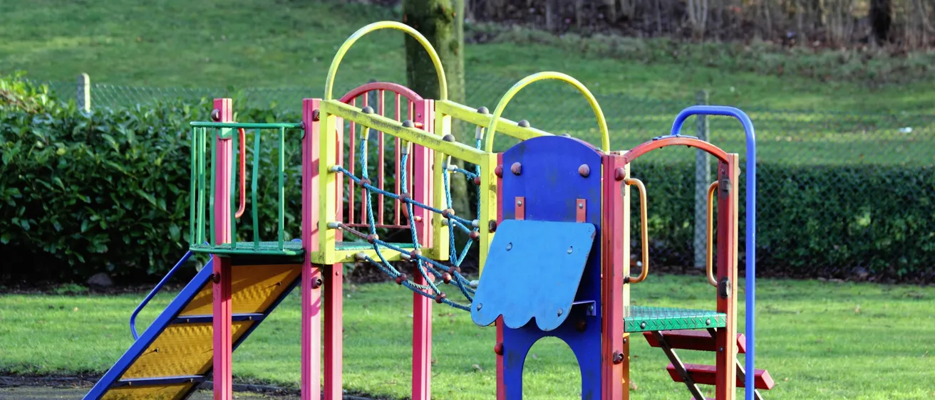 Ein farbenfroher Spielplatz mit Rutsche, Kletternetz und Treppen, auf einer Rasenfläche mit einer Hecke und Bäumen im Hintergrund.