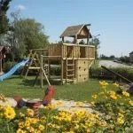 Ein Außenspielplatz mit einem hölzernen Klettergerüst, einer Rutsche und einem Rasenbereich mit gelben Blumen.