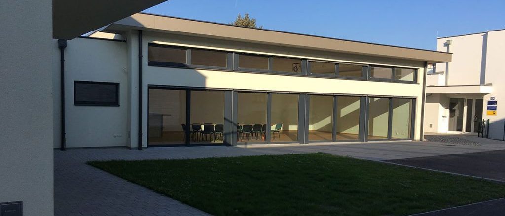 Ein modernes weißes Gebäude mit Glastüren und -fenstern befindet sich in einem Außenbereich. Es gibt Stühle im Gebäude.