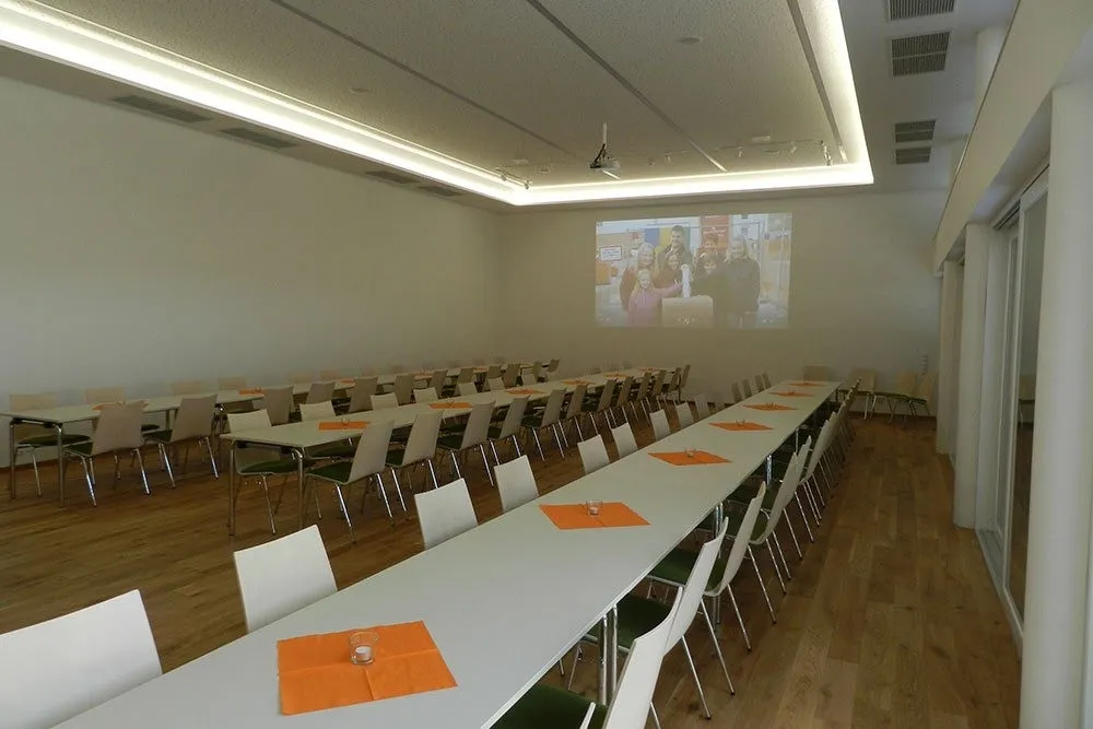 Ein Konferenzraum mit einer Projektionsleinwand, die ein Familienfoto zeigt. Tische und Stühle sind ordentlich angeordnet. Der Raum ist gut beleuchtet und leer.
