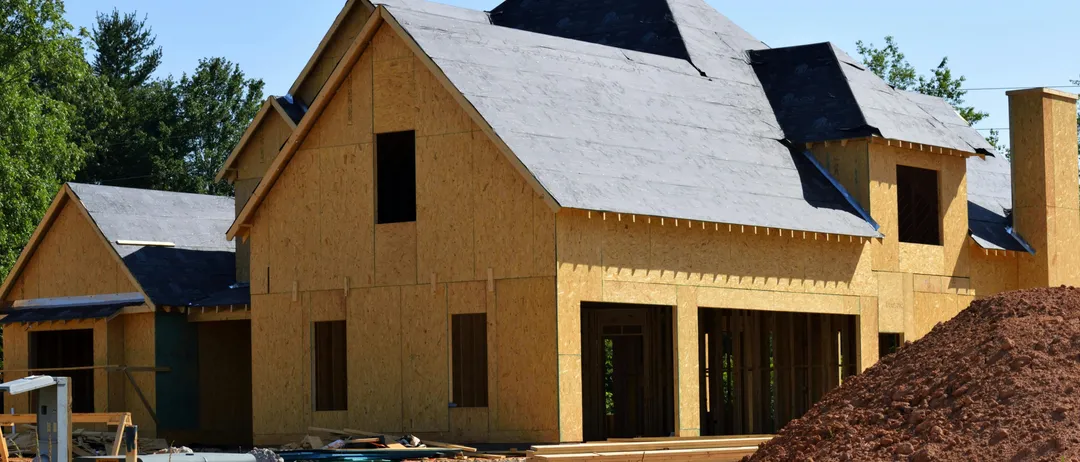 Ein Haus im Bau mit einem grauen Dach, hölzerne Außenwand und mehreren Fenstern und Türen. Es ist von Erde und Kies umgeben, mit einem Haufen Steine in der Nähe.