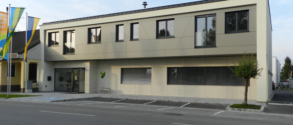 Ein Bürogebäude mit modernem Design steht vor einem Parkplatz, mit einem Baum und einem Fahnenmast. Das Gebäude hat einen verglasten Eingang und mehrere Fenster.