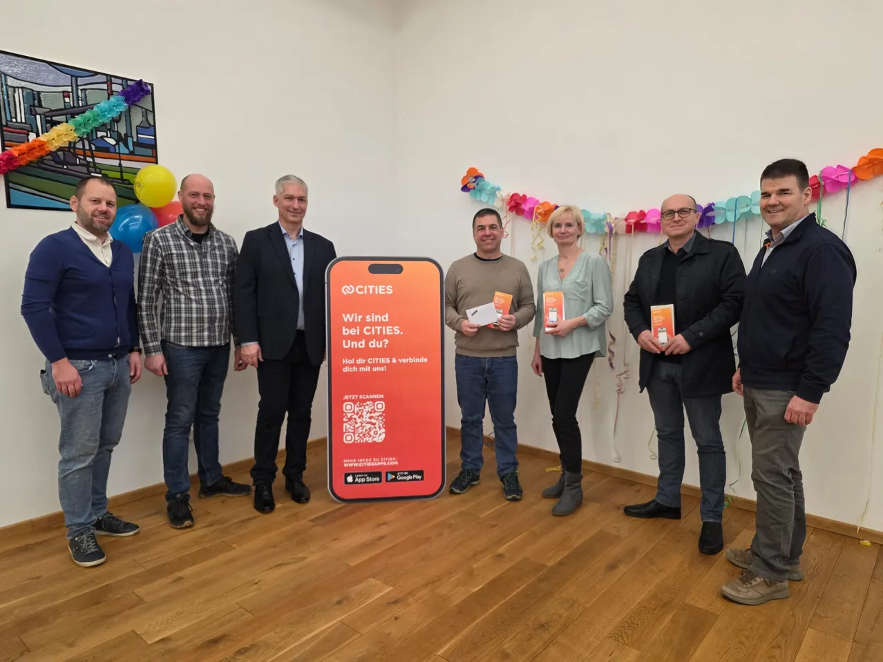 Eine Gruppe von Menschen steht neben einem roten Poster mit dem Text 'Wir sind CITIES. Und du?'. Sie halten Auszeichnungen und lächeln. Dahinter befindet sich eine bunte Girlande und eine Wand mit Luftballons.