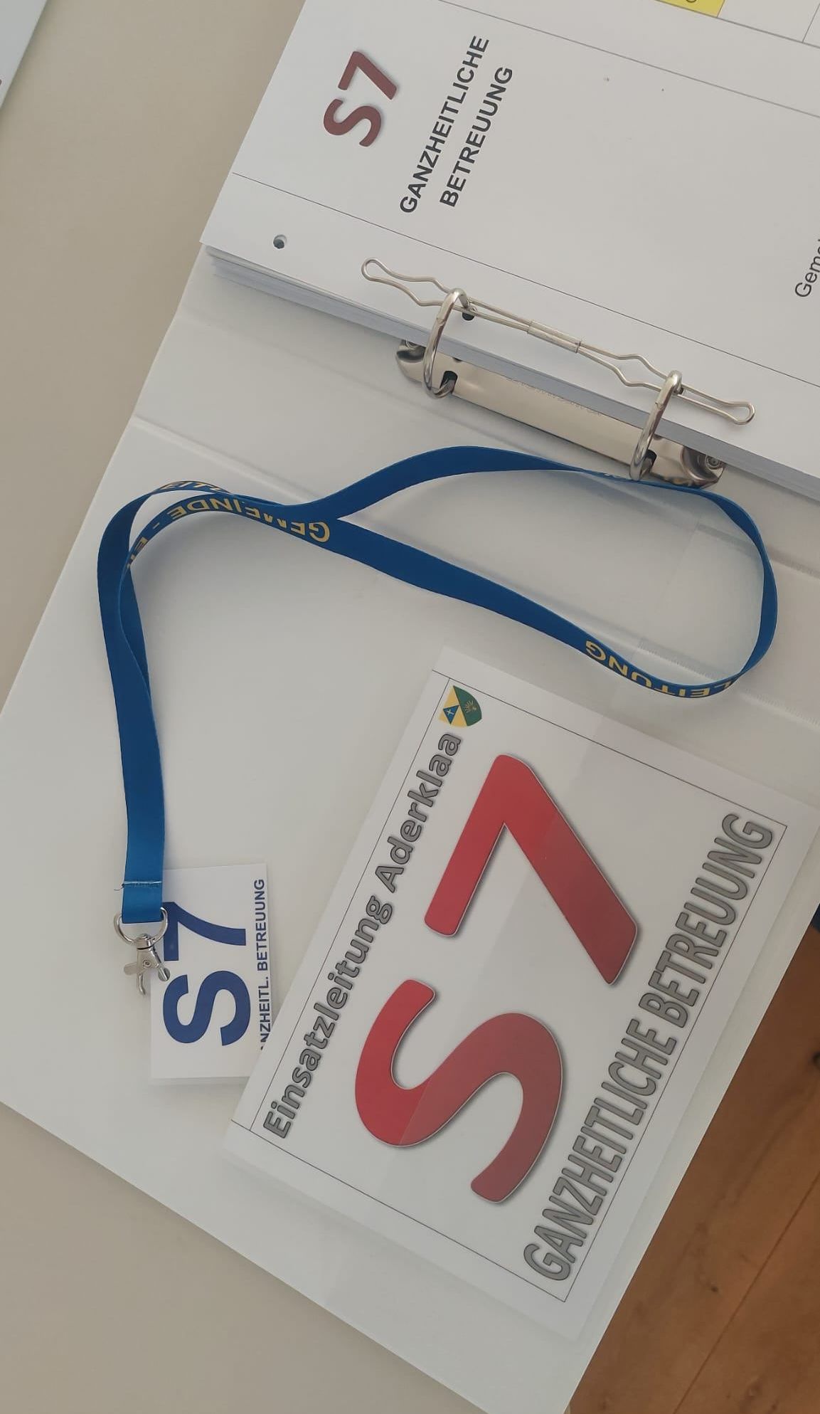 Ein Nahaufnahme eines blauen Lanyards mit der Nummer 57 darauf, auf einem weißen Ordner mit dem Text 'Ganzheitliche Betrachtung' und 'Betreuung'.