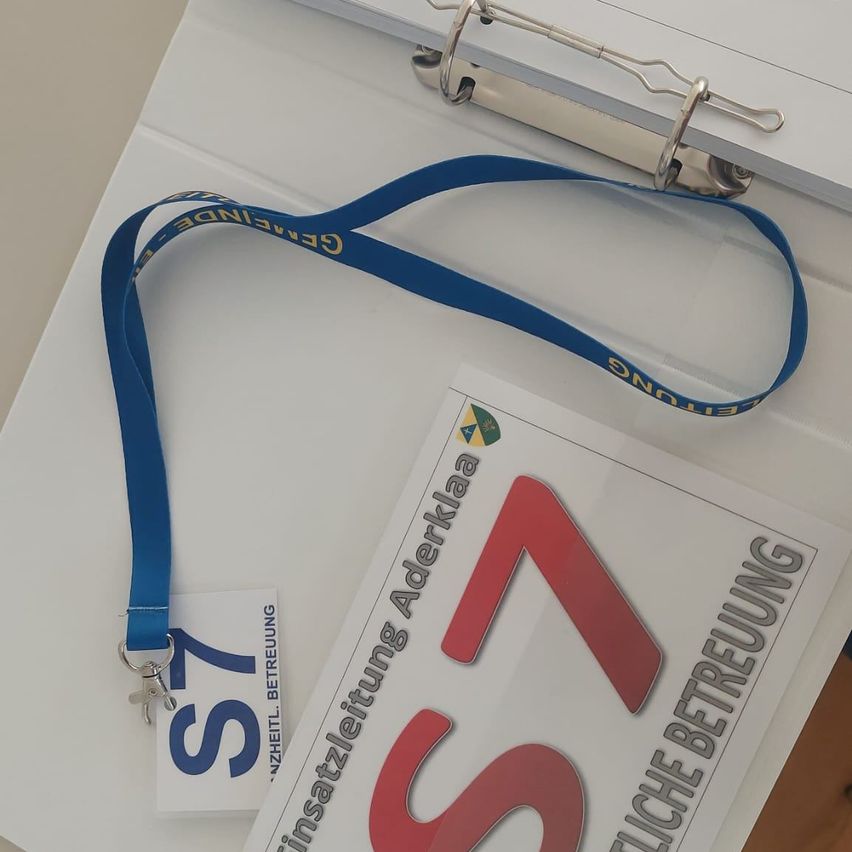 Ein Nahaufnahme eines blauen Lanyards mit der Nummer 57 darauf, auf einem weißen Ordner mit dem Text 'Ganzheitliche Betrachtung' und 'Betreuung'.