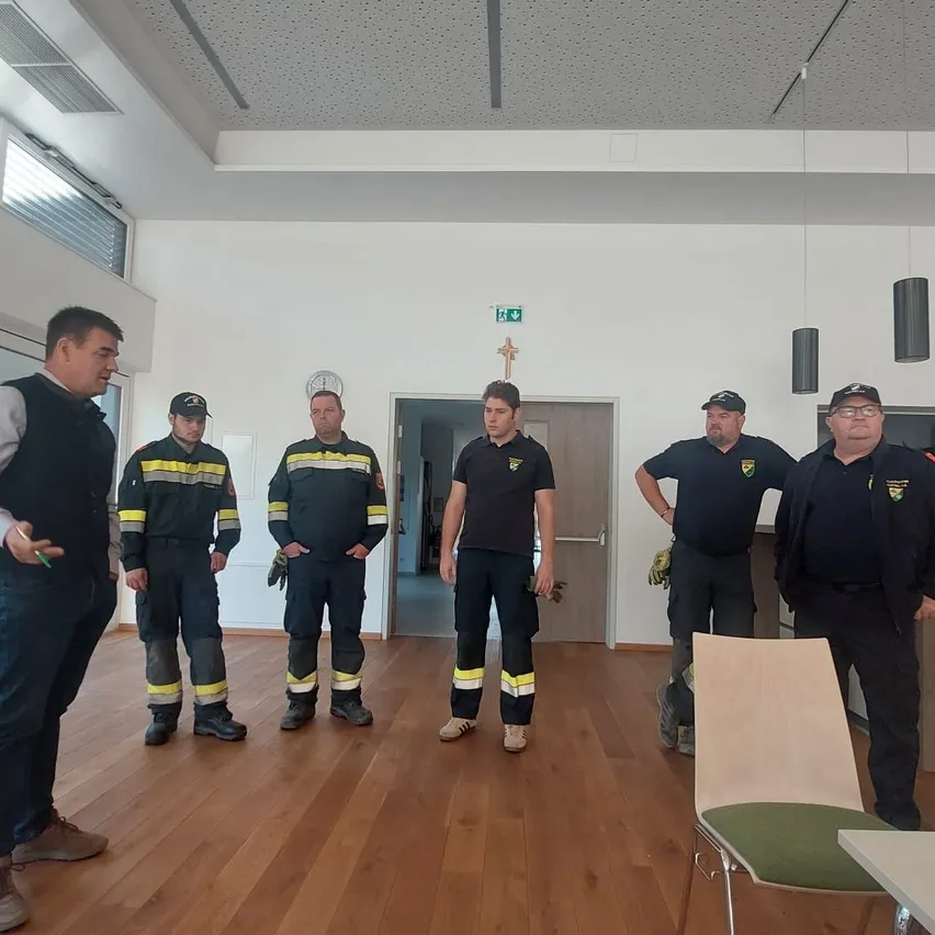 Eine Gruppe von Feuerwehrleuten steht in einem Raum, wobei ein Mann die anderen anspricht.