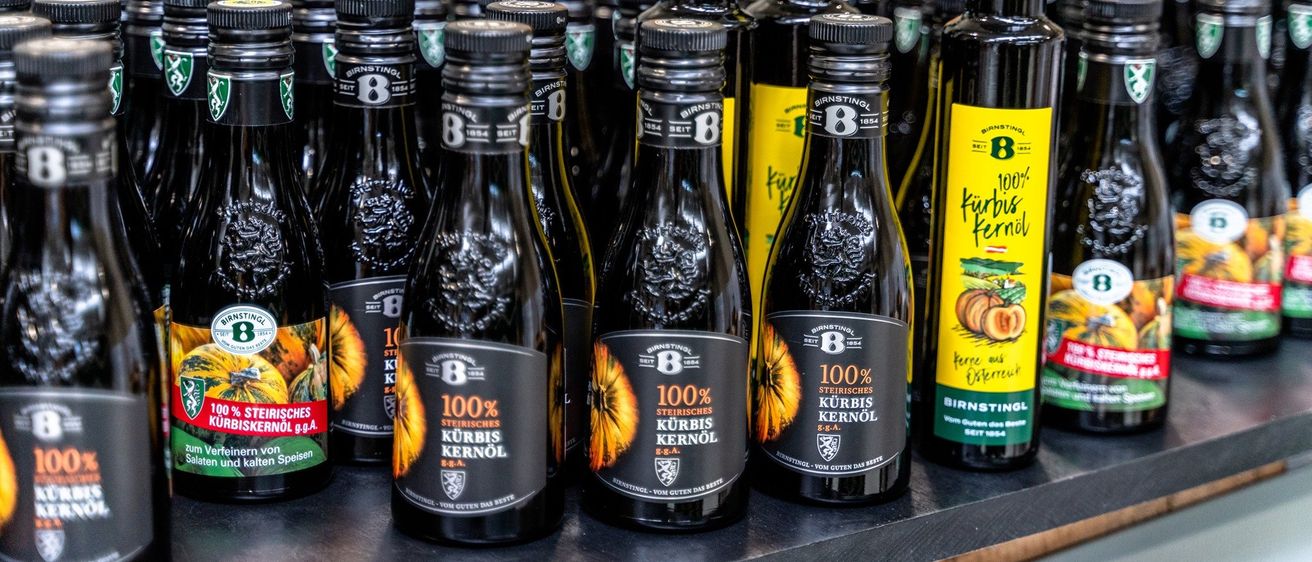 Eine Reihe von Flaschen gefüllt mit Kürbisöl, mit grünen und gelben Etiketten. Die Etiketten lesen '100% Kürbis Kernöl' und 'Kürbis öl', was darauf hindeutet, dass es sich um Kürbiskernöl handelt. Die Flaschen sind auf einem Regal angeordnet.