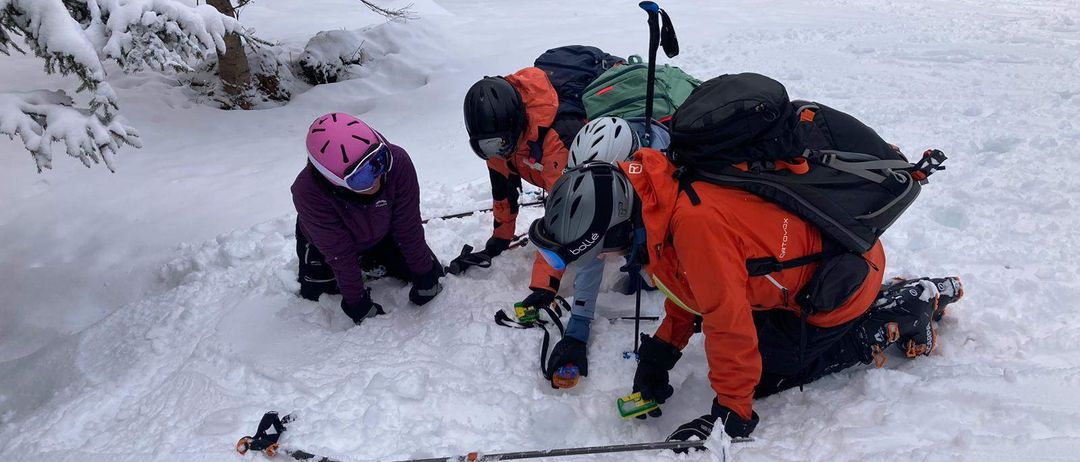 Eine Gruppe von Menschen mit Helmen und Rucksäcken skifahren im Schnee. Sie sind auf den Knien und schauen auf den Boden.