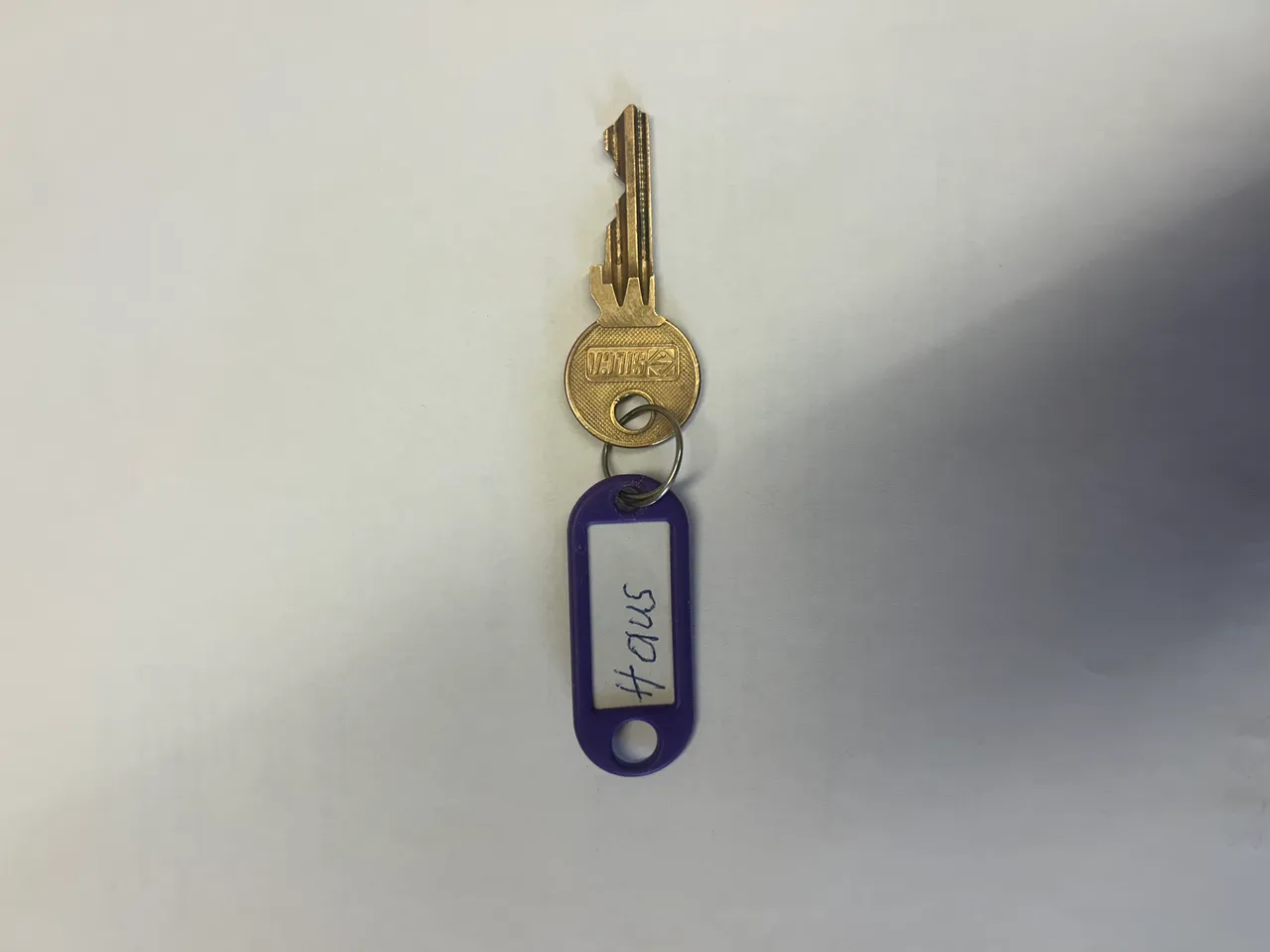 Ein goldener Schlüssel mit einem lila Schlüsselanhänger, auf dem 'HAUS 3164' steht, wird vor einem weißen Hintergrund gezeigt.