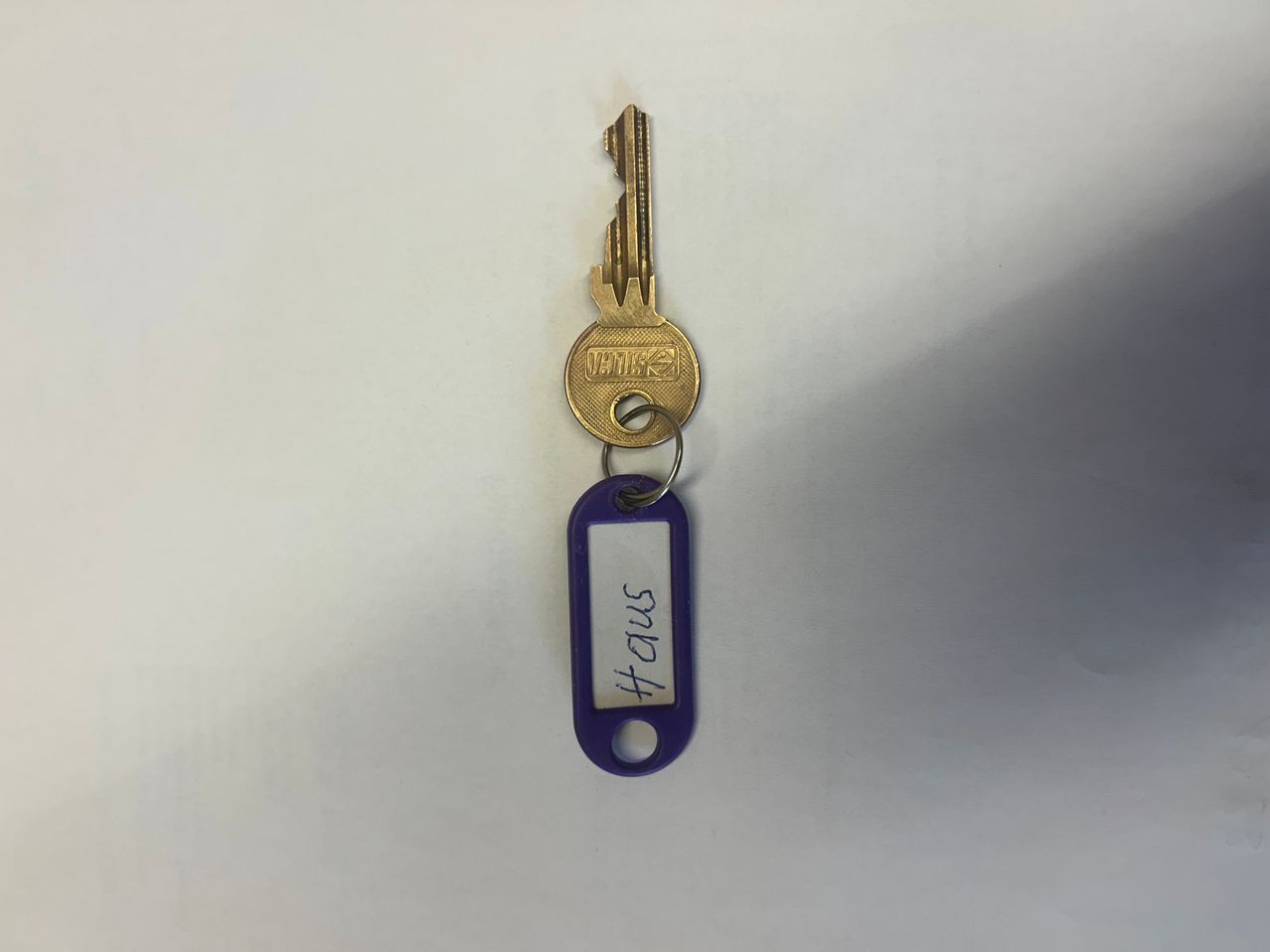 Ein goldener Schlüssel mit einem lila Schlüsselanhänger, auf dem 'HAUS 3164' steht, wird vor einem weißen Hintergrund gezeigt.