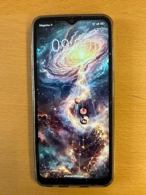 Ein Smartphone mit einem galaktischen Bild auf dem Bildschirm steht auf einem Holztisch. Es zeigt die Zeit 09:00 an, und das Datum ist der 24. Februar 2026.