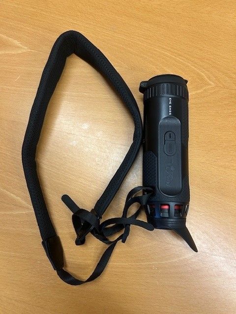 A black thermal imaging camera is attached to a black strap, placed on a wooden surface.