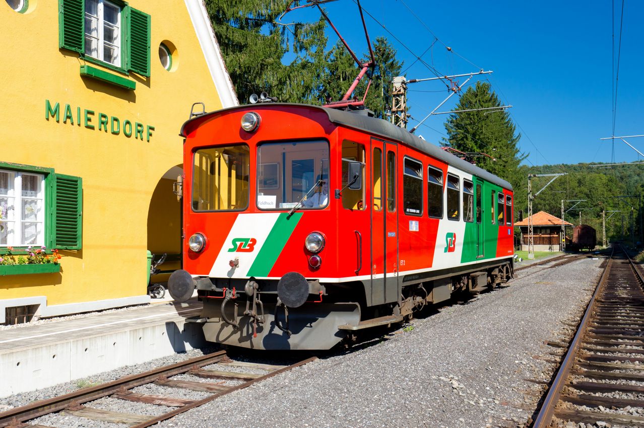Ein roter Zug mit grünen und weißen Streifen steht auf einem Schottergleis.