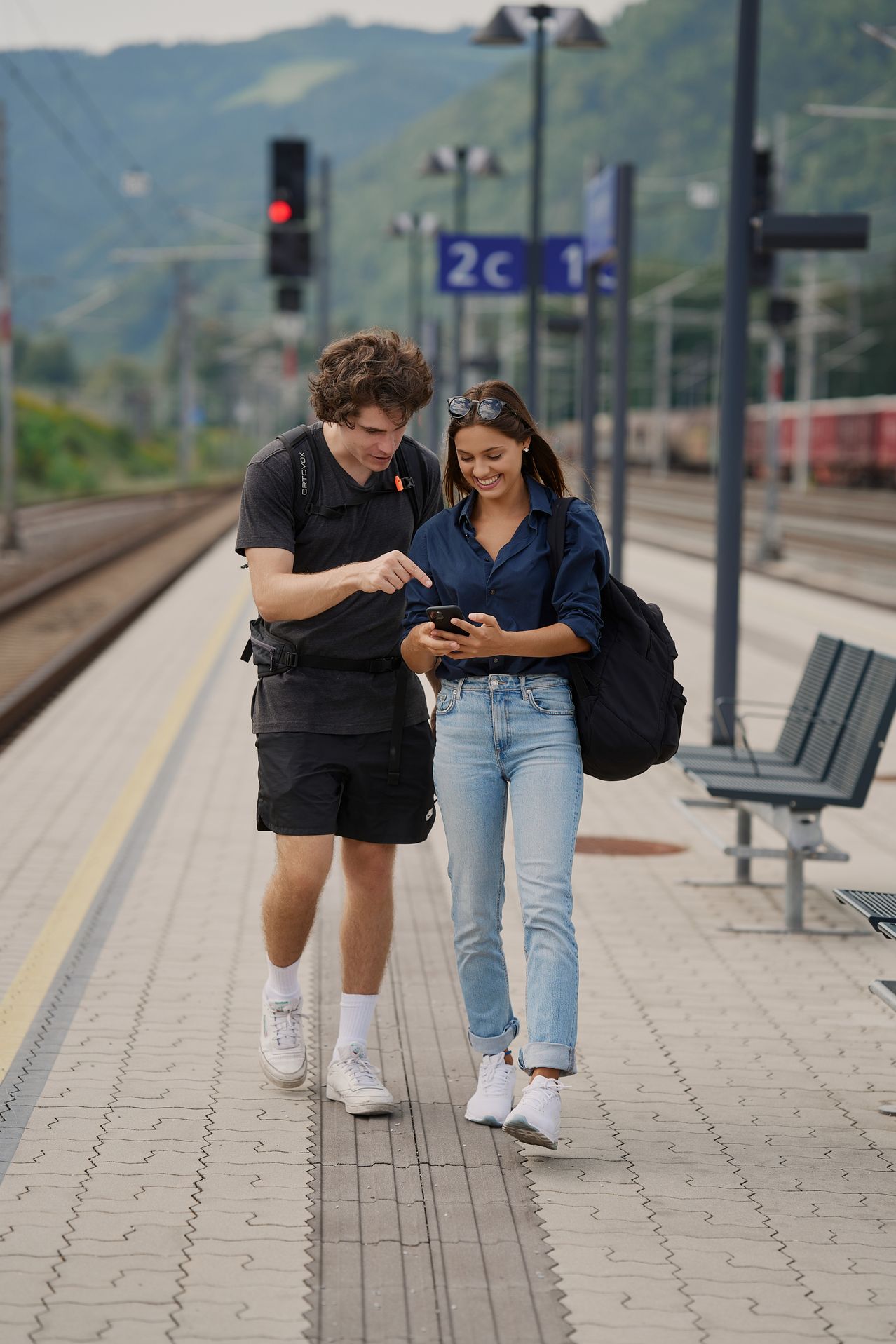 Ein Mann und eine Frau gehen auf einem Bahnsteig. Der Mann trägt einen Rucksack und schaut auf sein Handy, während die Frau lächelt und ebenfalls ihr Handy benutzt. Hinter ihnen befinden sich Metallpfosten, eine Bank und ein Zug.