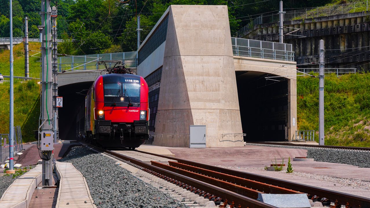 Ein roter Zug mit der Nummer 1116 235 fährt in einen Tunnel. Der Tunnel hat einen Betonpfeiler und eine Brücke darüber. Der Zug fährt auf den Gleisen mit Kies an der Seite.