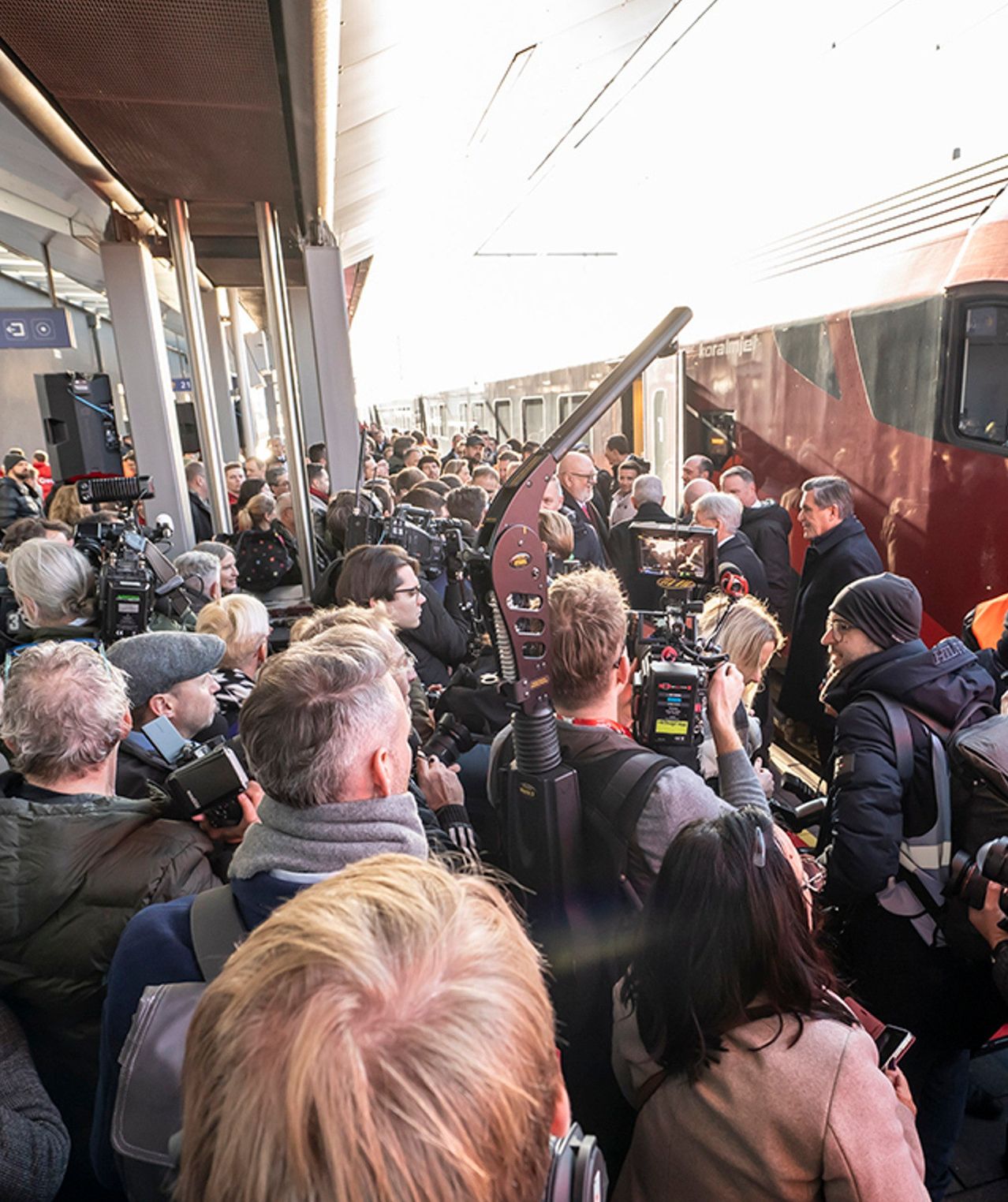 Eine Menschenmenge versammelt sich an einem Bahnhof und hält den Moment mit Kameras fest. Einige Personen tragen Mützen und Jacken, und der Zug ist im Hintergrund zu sehen.