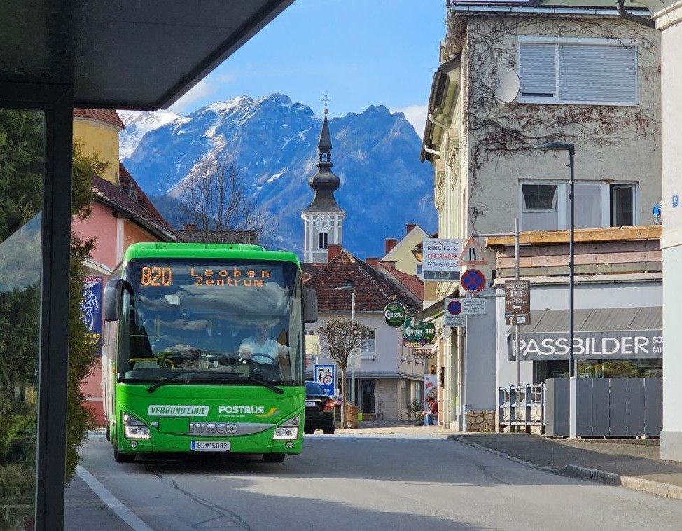 Ein grüner Bus mit der Aufschrift 'VERBUND LINE' und 'POSTBUS' fährt auf einer Straße mit Bergen im Hintergrund. Eine Person sitzt auf dem Gehweg in der Nähe einer Straßenlaterne.