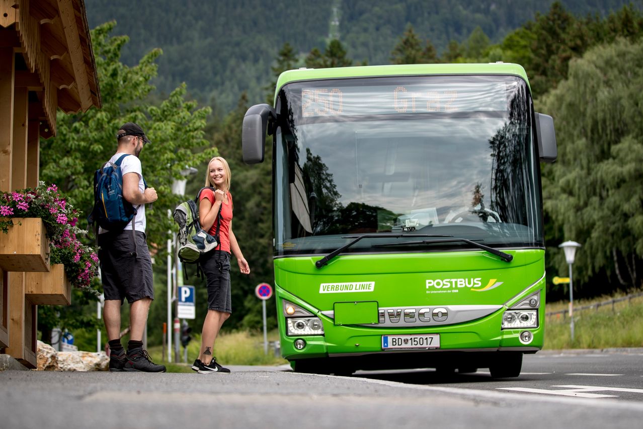 Eine Frau und zwei Männer stehen neben einem grünen Bus, mit Bergen und Bäumen im Hintergrund. Der Bus hat 'VERBUND LINIE' und 'POSTBUS' auf seiner Front.