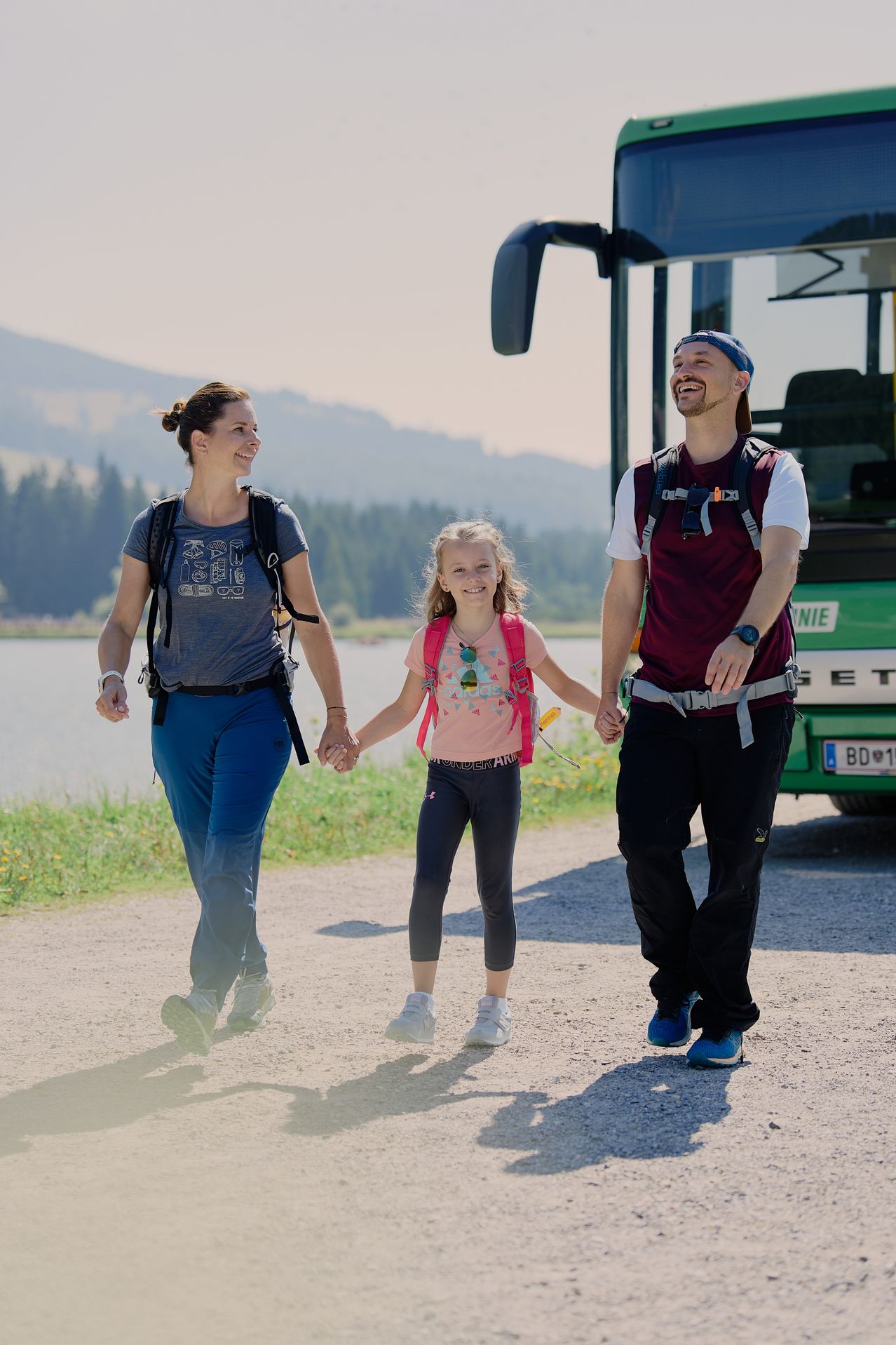 Eine dreiköpfige Familie läuft gemeinsam auf einem Pfad in der Nähe eines Sees, mit einem Bus hinter ihnen. Die Frau, der Mann und das Mädchen lächeln und halten Händchen.