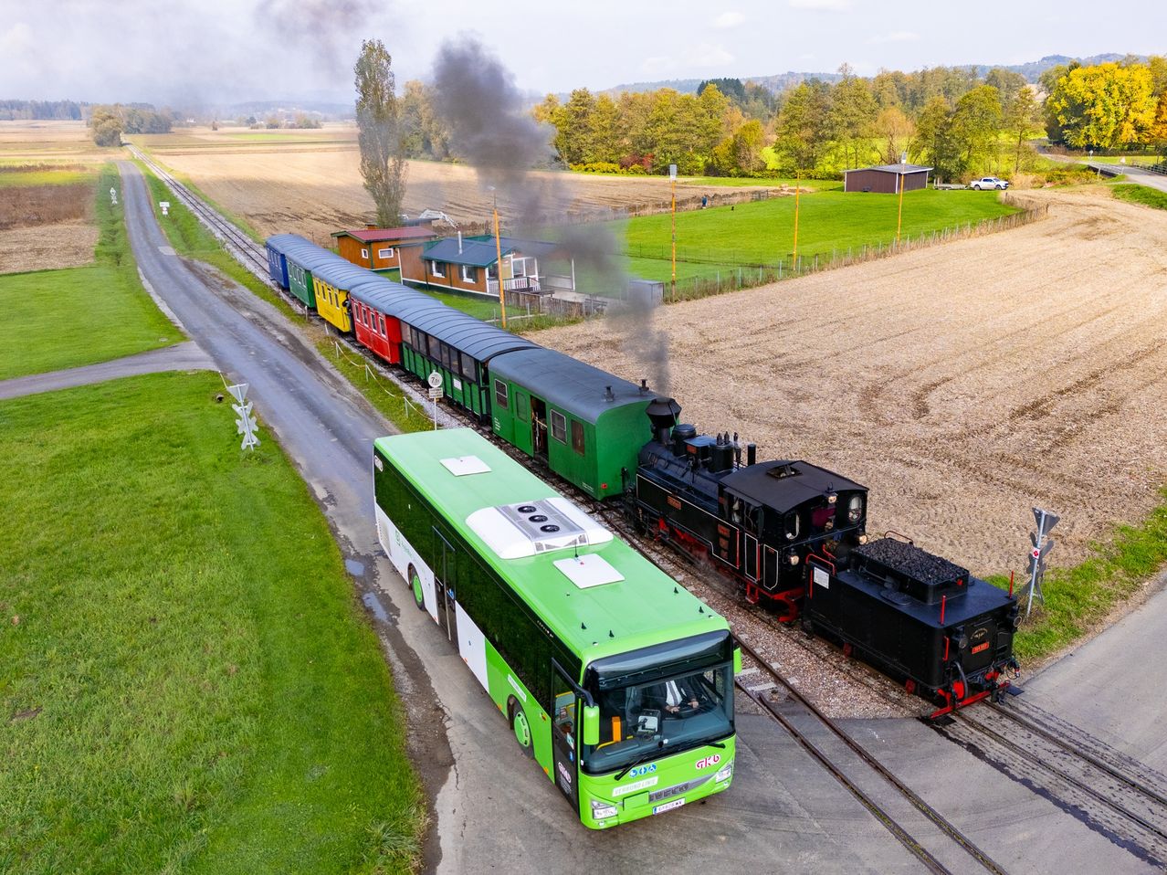 Ein bunter Zug fährt durch ein Feld auf einer Bahnstrecke. Ein grüner Bus steht neben ihm auf der Straße.