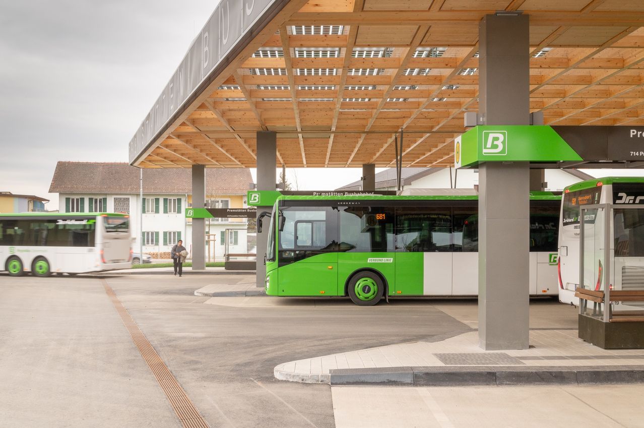 Ein grüner und weißer Bus hält an einer modernen Bushaltestelle mit Solarpanelen auf dem Dach. Eine Person geht in der Nähe mit einer Tasche.