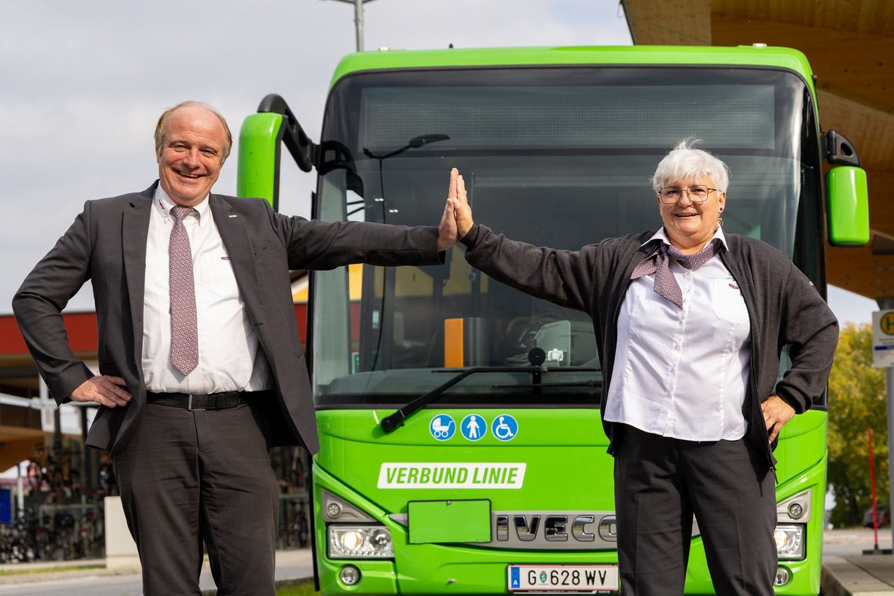 Zwei Personen stehen vor einem grünen Bus, sie berühren sich gegenseitig die Hände. Sie lächeln und scheinen für ein Foto zu posieren.