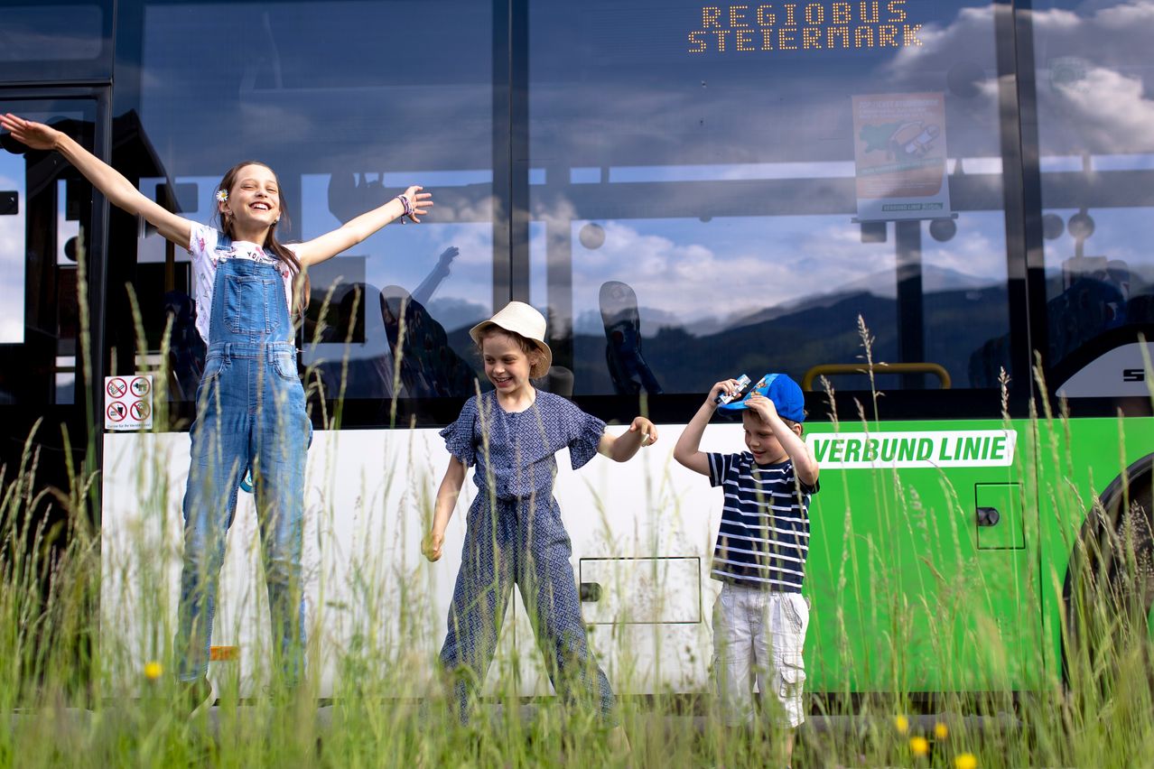 Drei Kinder stehen vor einem Bus auf einem Feld, lächeln und posieren für ein Foto. Das Mädchen hat die Arme erhoben, während die anderen zwei Mützen halten.